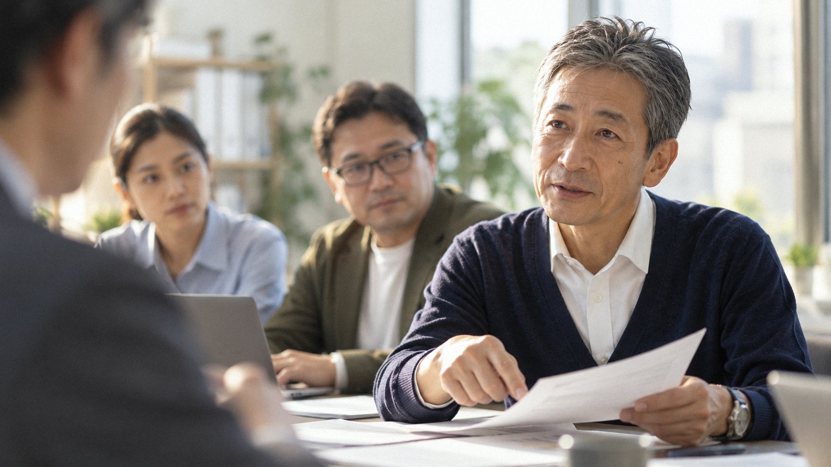 打合せ室で、所長の篠原が書類を手に相談者へ説明し、後ろで岩佐とグエン・ティ・ハーが聞いている